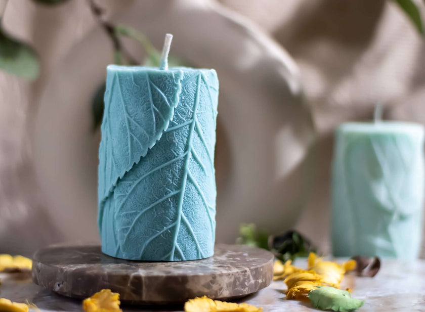 Handmade botanical decorative candle in light blue with leaf texture, placed on a marble base, surrounded by dried flowers.