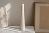 Tall white candle on a white surface with a framed abstract art piece in the background.