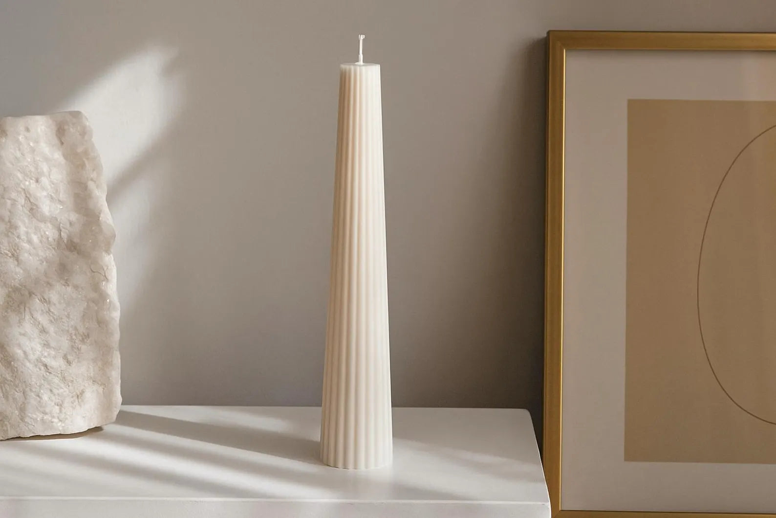Tall white candle on a white surface with a framed abstract art piece in the background.