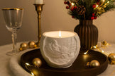 White embossed candle in a decorative setting with gold ornaments and a vase of flowers.