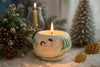 Decorative candle with festive design surrounded by pine cones and small trees