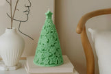 Green tree-shaped candle on a white surface with a vase and chair in the background.