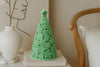 Green tree-shaped candle on a white surface with a vase and chair in the background.