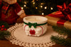 Decorative candle with a green bow on a doily, surrounded by Christmas decorations including a gift and tree.