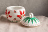 Decorative ceramic pot with red floral design and matching lid on a neutral background