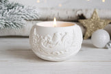 White ceramic candle holder with a lit candle on a wooden surface with decorative items in the background.