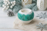 Decorative candle in a textured holder with winter-themed decorations on a light wooden surface.
