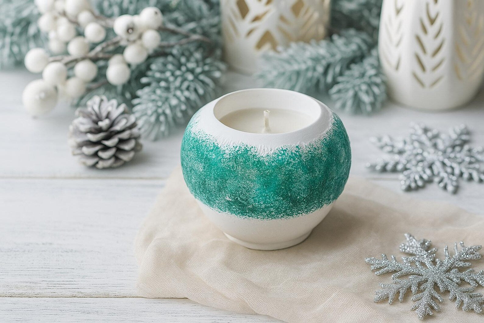 Decorative candle in a textured holder with winter-themed decorations on a light wooden surface.