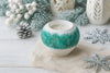 Decorative candle in a textured holder with winter-themed decorations on a light wooden surface.