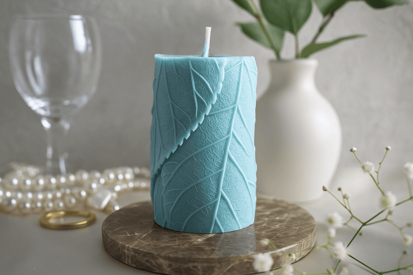 Teal candle with leaf pattern on a wooden stand, with a vase and glass in the background.