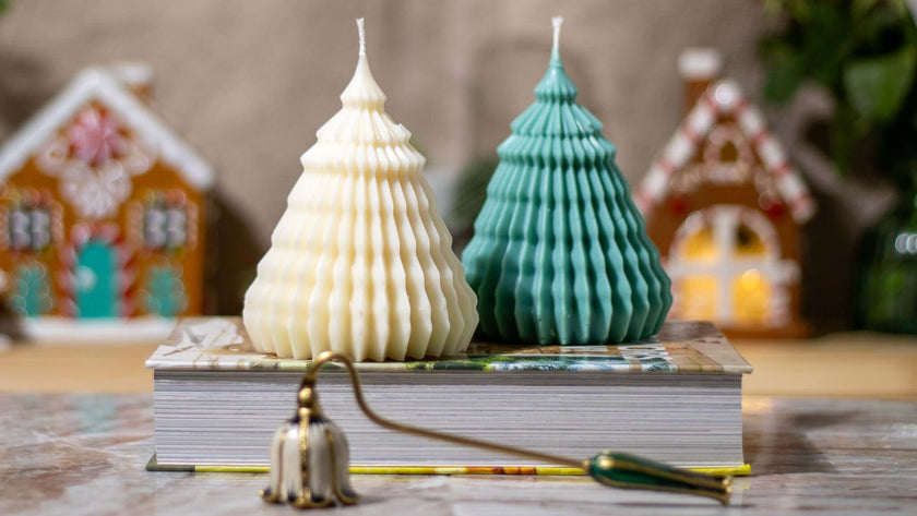 Two decorative tree-shaped candles, one cream and one teal, placed on a book with festive gingerbread houses in the background.