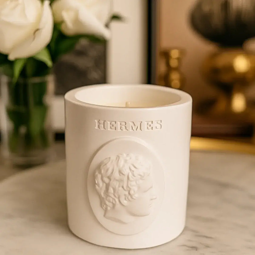 White Hermes candle on a marble surface with flowers in the background