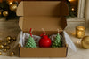 Three festive candles shaped like trees and a globe, and present in an open cardboard box.