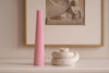 Pink candle on a white surface with a framed artwork in the background