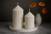 Two ribbed candles on a round ceramic holder with a vase of orange flowers in the background.