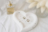 Heart-shaped ceramic dish with earrings on a white surface next to a perfume bottle.