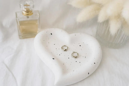 Heart-shaped ceramic dish with earrings on a white surface next to a perfume bottle.