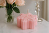 Pink snowflake-patterned candle on a marble surface with a vase of pink roses in the background.