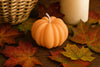 Pumpkin-shaped candle on a surface with autumn leaves and a woven basket in the background. Pumpkin shaped soy candle made with natural eco-friendly wax for fall home décor
