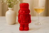 Red nutcracker-shaped candle on a table with a vase and glass in the background