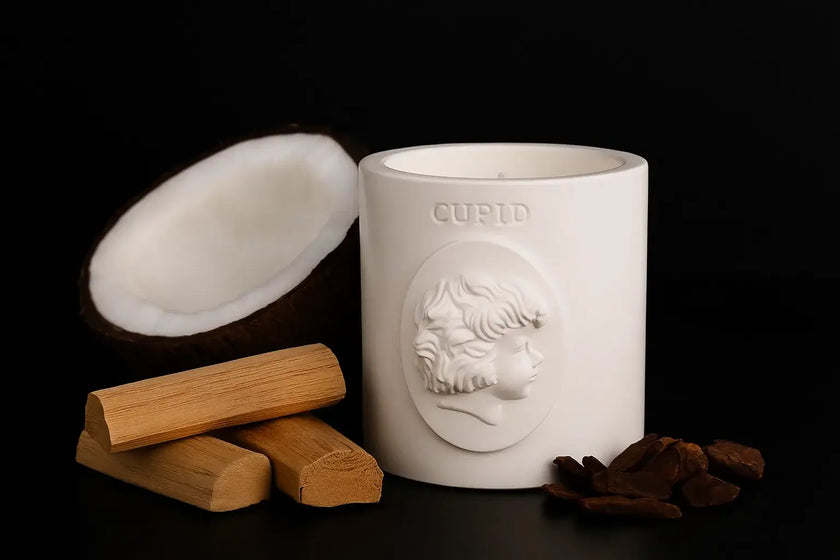 White candle with 'Cupid' design, wooden wick trimmers, and dried herbs on a black background