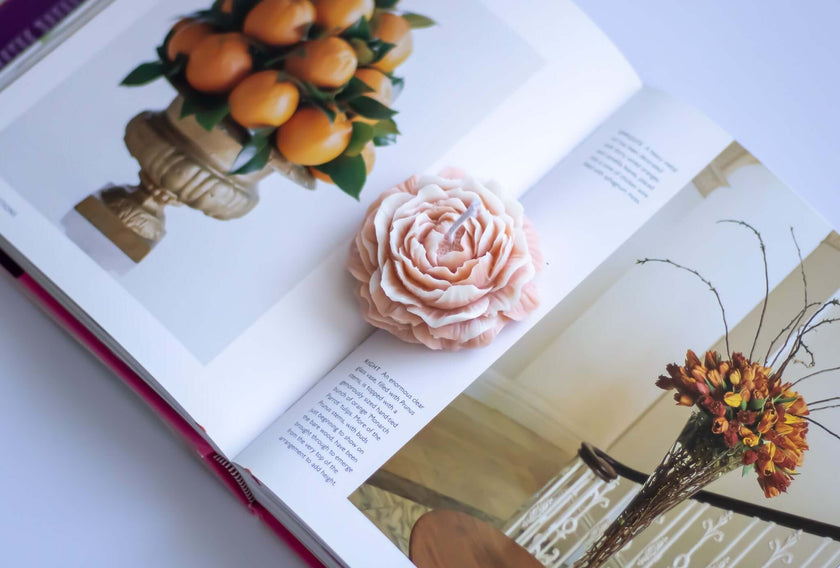 Rose Blossom candle on an open book, showcasing sculptural soy wax flower next to fruit and floral arrangements.
