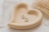 Heart-shaped beige dish with silver hoop earrings on a soft textured background
