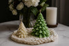 Two Christmas tree-shaped candles on a textured surface with flowers and a candle in the background.