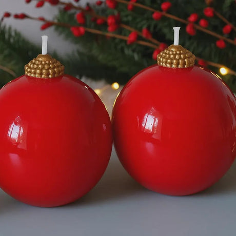 Two red ball candles with gold tops on a festive background with greenery and lights.