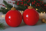 Two red ball candles with gold tops on a festive background with greenery and lights.