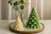 Two tree-shaped candles, one white and one green, on a marble tray with a vase of flowers in the background.