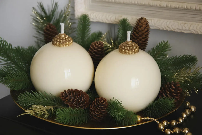 Two white spherical candles with gold tops on a bed of greenery and pine cones.