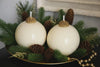 Two white spherical candles with gold tops on a bed of greenery and pine cones.