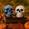 Two skull-shaped candles, one gray and one cream, displayed on top of a vintage book surrounded by autumn leaves and small pumpkins. Scented Gothic Skull Soy Wax Candle – Halloween Decor