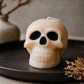 An off-white skull-shaped candle with black eye sockets and a white wick, displayed on a dark round tray with dried flowers and a blurred vase in the background. Scented Gothic Skull Soy Wax Candle – Halloween Decor