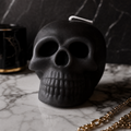 A matte black skull-shaped candle with a white wick, displayed on a white marble surface with a dark marble background. Scented Gothic Skull Soy Wax Candle – Halloween Decor