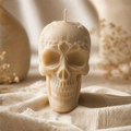A cream-colored skull-shaped candle with a single wick, standing upright on draped beige fabric with soft, blurred decorative elements in the background. Skull Soy Wax Candle – Gothic Halloween Decor