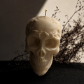 An off-white skull-shaped candle with a small wick on top, set against a dark background with dried branches. Skull Soy Wax Candle – Gothic Halloween Decor