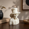 A white skeleton candle shaped like a man's bust wearing a top hat with a rose detail, lit and placed on a wooden table in a styled interior setting. Handmade Halloween skeleton candle for haunted home decor
