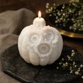 A white carved candle shaped like a pumpkin with three large flower details on the front, lit with a small flame, displayed on a dark stone surface with neutral fabric and small white flowers in the background. Decorative pumpkin candle for cozy table styling
