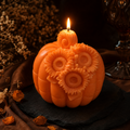 An orange pumpkin-shaped candle with carved sunflower details and a lit flame, displayed on a dark slate surface with warm autumn props in the background. Floral pumpkin soy wax candle for Thanksgiving decor
