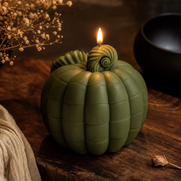 A sculpted pumpkin-shaped candle in olive green sits lit on a dark wooden surface, with a small flame at the top and dried floral props around it. Pumpkin soy wax candle for fall decor