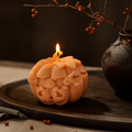 An orange carved pumpkin-shaped candle with a lit flame, styled on a dark wooden tray with a dark ceramic vase and dried branches in the background. Jack O Lantern pumpkin soy wax candle for Halloween decor