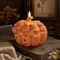 An orange carved pumpkin-shaped candle with a lit flame, styled on a rustic wooden surface with autumn-themed props in a warm, dimly lit setting. Handmade Halloween pumpkin candle with ghost details