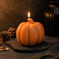 An orange pumpkin-shaped candle with a lit flame, displayed on a dark slate coaster with pinecones and a dark textured vase in the background. Cute spooky pumpkin candle for fall table styling