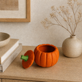 A small orange pumpkin-shaped container with a matching lid and a green stem detail sits on a wooden table in a styled interior setting, next to stacked books and a beige vase with dried branches.