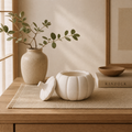 A white ribbed pumpkin-shaped ceramic container with its lid placed beside it on a woven beige mat, set on a wooden table in a softly lit interior. A speckled beige vase with green stems and a book labeled KINFOLK are also visible in the scene.