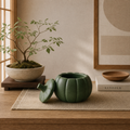 A green pumpkin-shaped ceramic container with its lid leaning beside it on a woven placemat on a wooden table. The scene includes a potted bonsai tree, a framed abstract print, a small wooden bowl, and a book labeled KINFOLK in a softly lit interior setting.