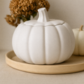 A white ceramic pumpkin-shaped container with a lid and stem, displayed on a light wood tray with dried flowers in the background.