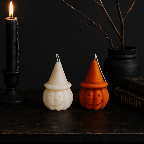 Two Halloween-themed wax candles on a dark wooden surface, one shaped like a cream-colored witch hat pumpkin and the other like an orange jack-o'-lantern with a pointed hat, styled with a lit black candle, a black cauldron, and books in the background. Pumpkin Hat Soy Wax Candles